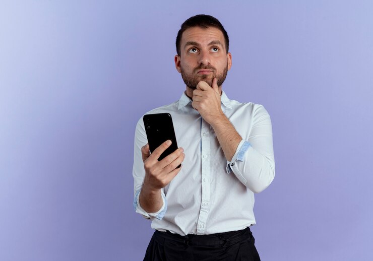 Foto grátis homem bonito pensativo coloca a mão no queixo segurando o telefone, olhando isolado na parede roxa