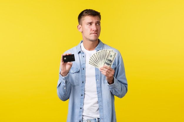 Homem bonito pensativo e indeciso, preocupado em roupas casuais, parecendo perplexo e mordendo o lábio, tentando comprar algo caro, mostrando um cartão de crédito com dinheiro.