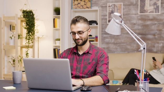 Homem bonito rindo enquanto trabalhava no laptop na sala de estar. Namorada em segundo plano fala ao telefone.