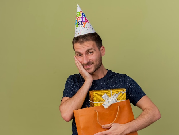 Homem bonito satisfeito com boné de aniversário coloca a mão no rosto e segura a caixa de presente em uma sacola de papel, isolada na parede verde oliva com espaço de cópia