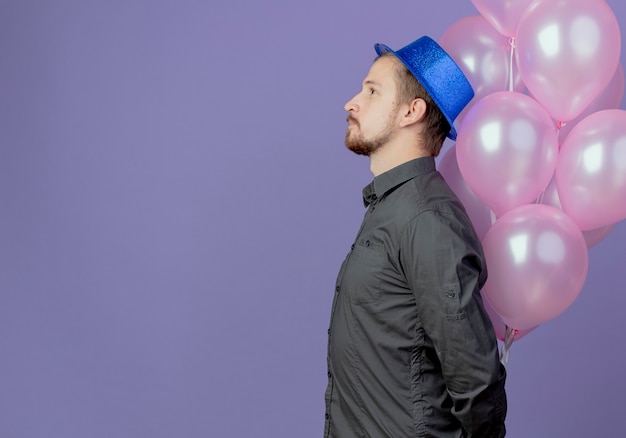 Homem bonito satisfeito com chapéu azul parado de lado com balões de hélio isolados na parede roxa