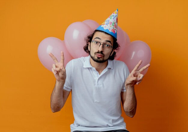 Homem bonito satisfeito usando óculos e boné de aniversário em pé na frente de balões e mostrando um gesto de paz isolado em um fundo laranja