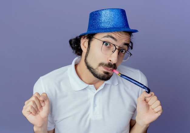 Homem bonito satisfeito usando óculos e chapéu azul, soprando apito e mostrando um gesto de sim