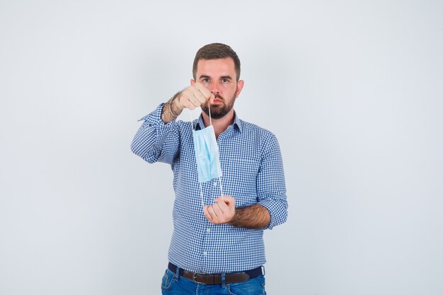 Homem bonito, segurando a máscara com as duas mãos na camisa, jeans e olhando sério, vista frontal.