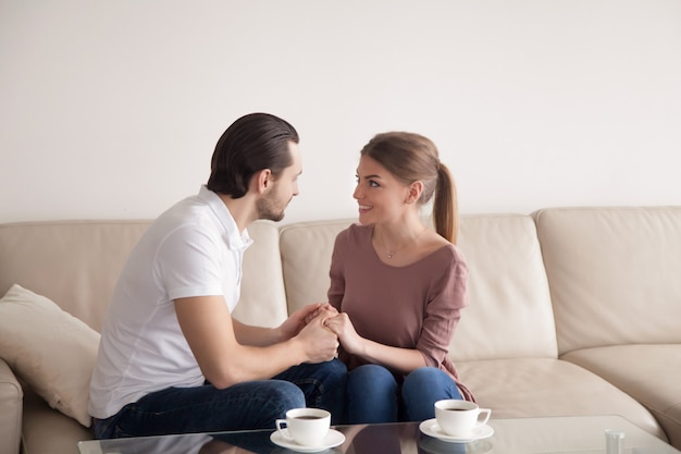 Homem bonito, segurando as mãos de mulher bonita sentada dentro de casa, proposta