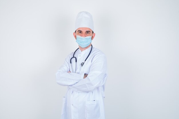 Homem bonito, segurando o estetoscópio em volta do pescoço, os braços em pé cruzados no jaleco branco de laboratório médico, máscara e parecendo confiante. vista frontal.