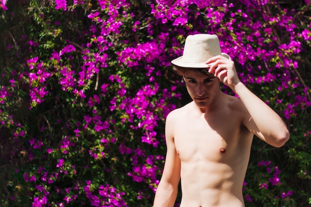 Homem bonito sem camisa, segurando o chapéu enquanto pé flor árvore no exterior