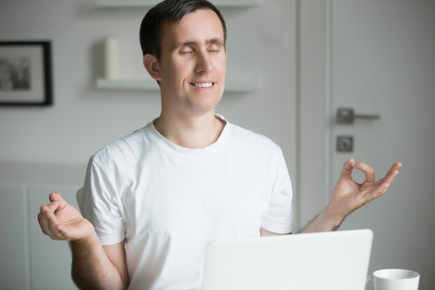 Homem bonito sentado em pose de yoga perto da mesa com laptop