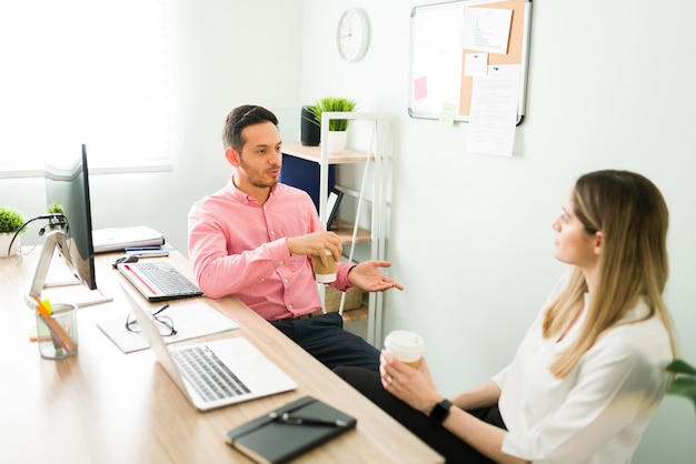 Homem bonito sentado em sua mesa e conversando com uma colega de trabalho e parceira de negócios sobre um acordo com um cliente. Colegas tomando café no escritório