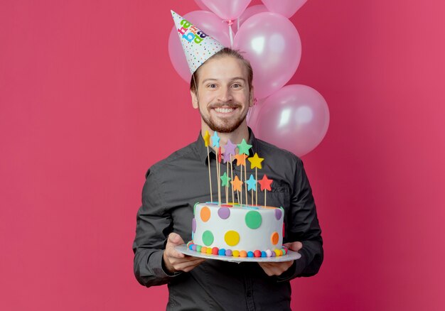 Foto grátis homem bonito sorridente com boné de aniversário parado com balões de hélio segurando um bolo de aniversário