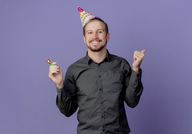 Homem bonito sorridente com boné de aniversário segurando apito e polegar para cima isolado na parede roxa