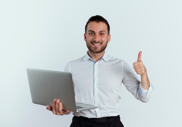 Homem bonito sorridente segurando laptop com polegar para cima isolado na parede branca