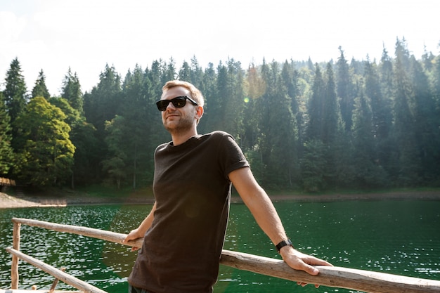 Homem bonito, sorrindo, apreciando a vista das montanhas, lago e floresta