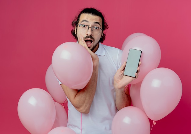 Homem bonito surpreso de óculos, em pé entre balões, segurando o telefone e colocando a mão na bochecha isolada na parede rosa
