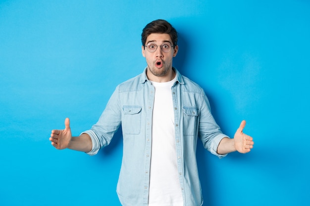 Homem bonito surpreso segurando algo grande, mostrando tamanho grande com rosto surpreso, em pé sobre um fundo azul