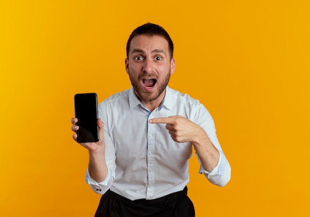 Homem bonito surpreso segurando e apontando para o telefone, parecendo isolado na parede laranja