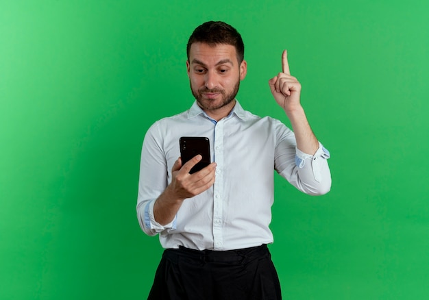 Homem bonito surpreso segurando e olhando para o telefone apontando para cima, isolado na parede verde