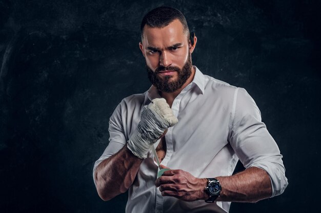 Homem bravo brutal na camisa branca está usando proteção no punho antes da luta no estúdio escuro.