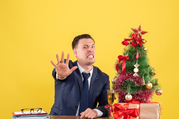 Homem bravo de frente, fazendo sinal de pare, sentado à mesa perto da árvore de natal e presentes em fundo amarelo