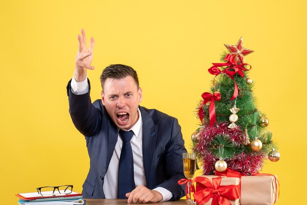 Homem bravo de frente, gritando enquanto está sentado à mesa perto da árvore de natal e presentes em fundo amarelo