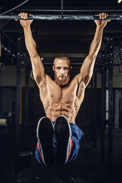 Homem brutal musculoso com barba fazendo pull up na barra horizontal em uma academia.