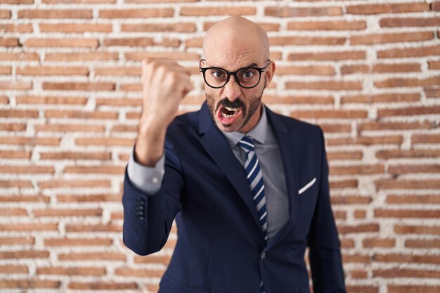 Homem careca com barba vestindo roupas de negócios e óculos bravo e louco levantando o punho frustrado e furioso enquanto gritava com raiva raiva e conceito agressivo