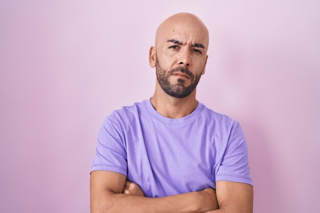 Homem careca de meia-idade em pé sobre fundo rosa cético e expressão de desaprovação nervosa no rosto com pessoa negativa de braços cruzados