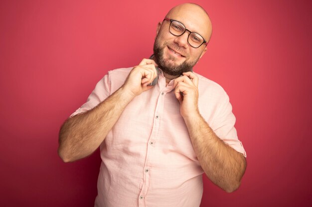 Homem careca de meia-idade, sorrindo, inclinando a cabeça e vestindo uma camiseta rosa com óculos e fones de ouvido no pescoço, isolados na parede rosa