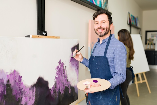 Foto grátis homem caucasiano atraente sorrindo e segurando um pincel enquanto está na frente da tela que ele está começando a pintar para uma aula de arte