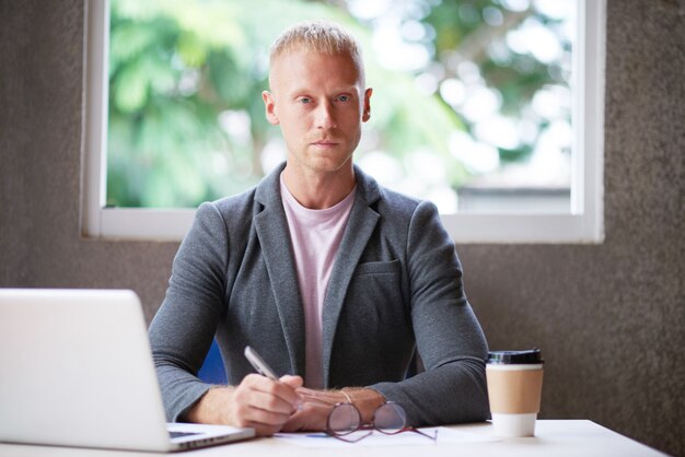 Homem caucasiano, blazer, sentando escrivaninha, em, escritório, com, laptop, e, olhando câmera