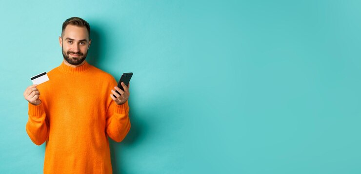 Foto grátis homem caucasiano bonito de compras on-line em suéter laranja usando cartão de crédito e telefone celular em pé