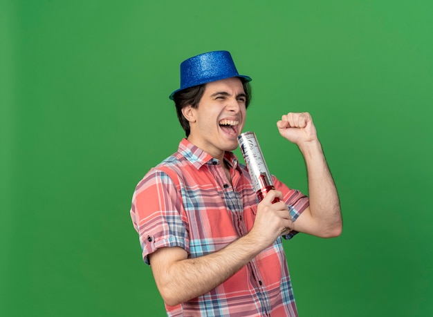 Foto grátis homem caucasiano bonito e alegre com chapéu de festa azul segurando um canhão de confete e segurando o punho