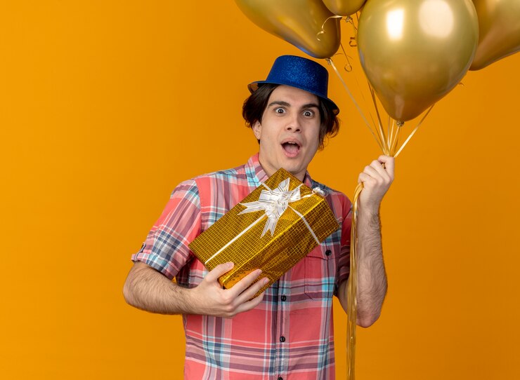 Foto grátis homem caucasiano bonito e animado com chapéu de festa azul segurando balões de hélio e uma caixa de presente