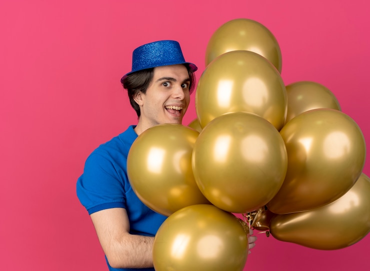 Foto grátis homem caucasiano bonito e animado com chapéu de festa azul segurando balões de hélio