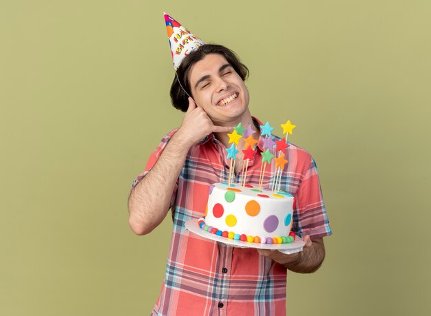 Homem caucasiano bonito e satisfeito com um boné de aniversário segurando um bolo de aniversário e gestos me chamam de sinal