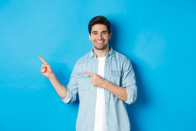 Homem caucasiano bonito em roupa casual, apontando o dedo para a esquerda e sorrindo, mostrando a oferta promocional, de pé sobre um fundo azul