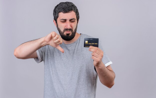 Homem caucasiano bonito jovem e desagradável mostrando e olhando para o cartão de crédito e mostrando o polegar para baixo, isolado no fundo branco com espaço de cópia