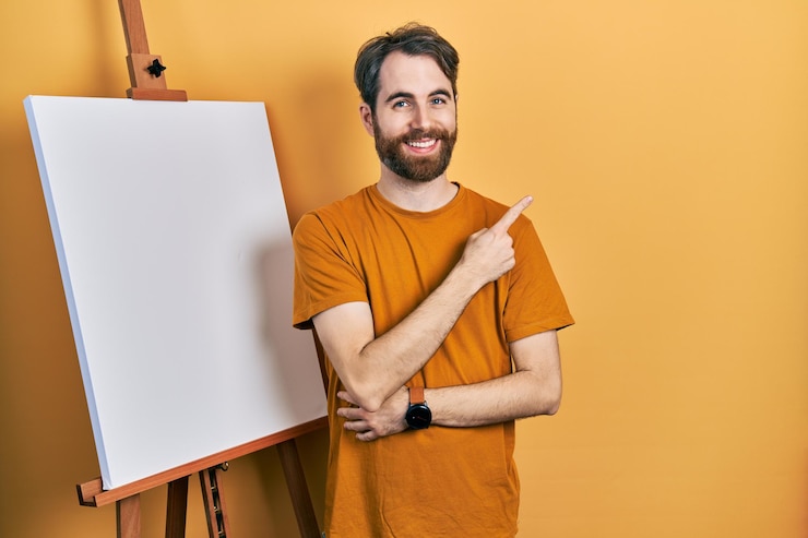 Foto grátis homem caucasiano com barba em pé ao lado do cavalete do pintor sorrindo alegremente apontando com a mão e o dedo para o lado