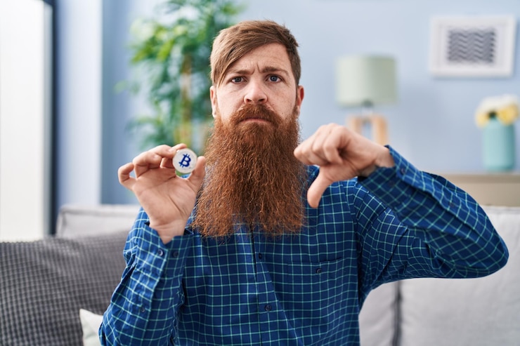 Foto grátis homem caucasiano com barba longa segurando bitcoin de moeda virtual com sinal negativo de rosto zangado, mostrando desagrado com o conceito de rejeição de polegares para baixo
