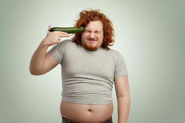 Homem caucasiano com cabelo ruivo usando uma camiseta pequena segurando pepino na têmpora como uma arma