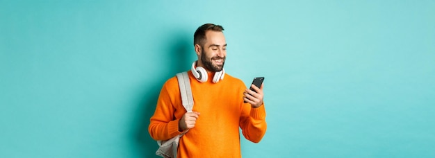 Foto grátis homem caucasiano com fones de ouvido e mochila olhando para o telefone lendo a mensagem e sorrindo em pé