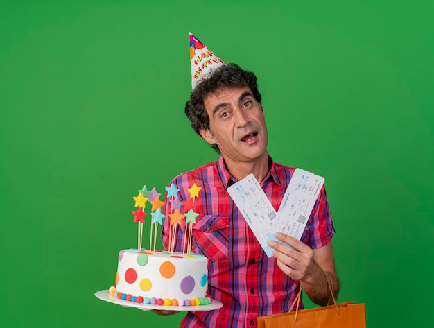 Homem caucasiano de meia-idade, festeiro, impressionado, usando um boné de aniversário, segurando um saco de papel de bolo de aniversário e passagens de avião, olhando para a câmera isolada no fundo verde com espaço de cópia