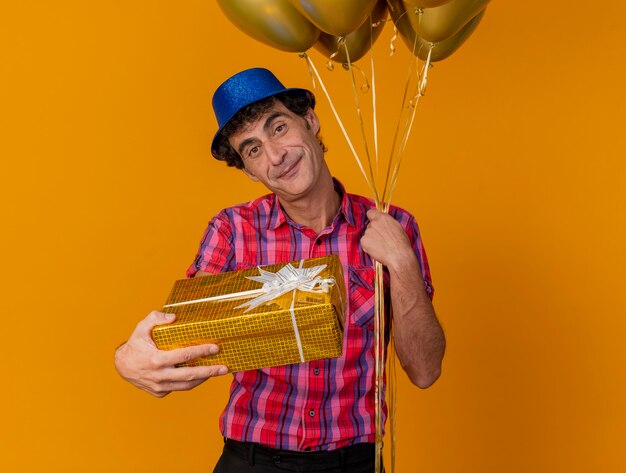 Homem caucasiano de meia-idade satisfeito com um chapéu de festa e segurando balões, olhando para a câmera estendendo o pacote de presente em direção à câmera