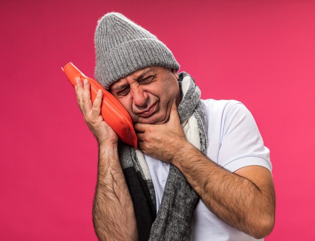 Homem caucasiano doente adulto desagradável com lenço no pescoço e chapéu de inverno coloca a mão no queixo e segura a garrafa de água quente isolada na parede rosa com espaço de cópia