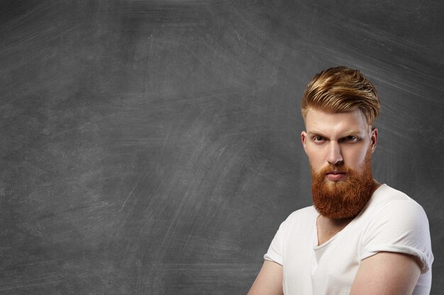 Homem caucasiano elegante com barba ruiva espessa e corte de cabelo moderno, posando com expressão séria e brutal no rosto no canto inferior direito contra um quadro-negro em branco com espaço de cópia para o seu conteúdo