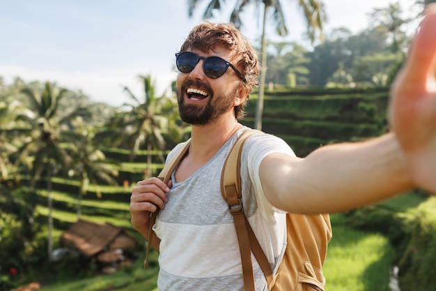 Homem caucasiano elegante feliz com mochila viajando na plantação de arroz e fazendo auto-retrato para as memórias.