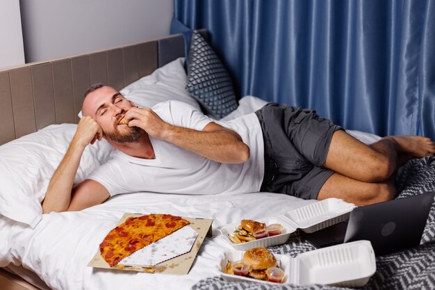 Homem caucasiano feliz comendo fast food em casa, no quarto na cama. O homem pediu comida online para levar e comeu pizza e hambúrgueres no quarto confortável