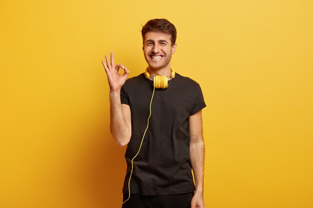 Homem caucasiano feliz mostra um gesto bom, concorda com algo, afirma que está tudo bem, sorri amplamente, usa fones de ouvido no pescoço