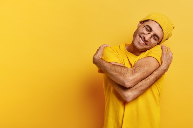 Homem caucasiano satisfeito se abraça, tem alta auto-estima, inclina a cabeça, tem um sorriso cheio de dentes, usa um chapéu amarelo casual e camiseta