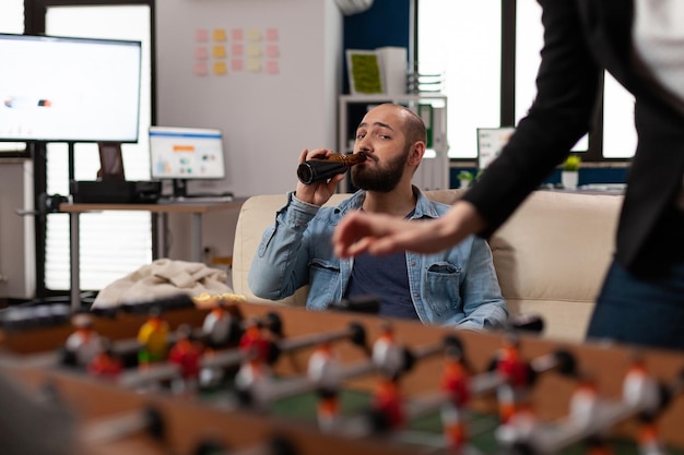 Homem caucasiano segurando garrafa de cerveja e assistindo jogo de pebolim, desfrutando de bebidas depois do trabalho com amigos. jovem comemorando com álcool, lanches, pizza, reunião com colegas de trabalho.
